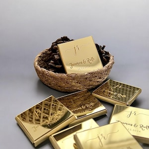 May include: Gold-coloured rectangular compact mirrors with engraved text, including initials and a date. Some mirrors are arranged around a woven basket filled with pinecones. The text reads "Jeremias & Ruth 26.12.2024".