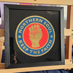 Puede incluir: Obra de arte enmarcada con borde negro. La obra presenta un diseño circular con las palabras "NORTHERN SOUL" y "KEEP THE FAITH" en letras doradas. Un círculo rojo en el centro contiene un puño dorado.