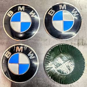 Op de afbeelding: Vier ronde zwarte en zilveren auto-emblemen met de letters BMW in wit en een blauw-wit propellerlogo in het midden. Een embleem is groen.
