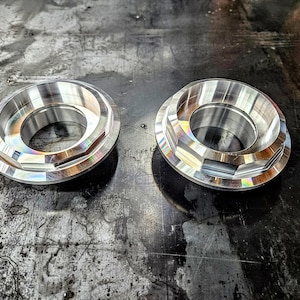 Puede incluir: Dos anillos metálicos mecanizados con un borde exterior facetado y un agujero central. Los anillos son de color plateado y reflejan la luz, creando un efecto arcoíris. Se colocan sobre una superficie oscura y texturizada.