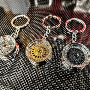 May include: Three silver keychains with different colored wheel designs. The keychains feature a silver ring, a silver chain, and a round wheel charm. The wheels are silver, gold, and black.