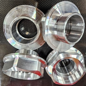 Puede incluir: Cuatro piezas metálicas plateadas con formas circulares y hexagonales. Dos piezas tienen aberturas roscadas y las otras aberturas lisas. Las piezas están dispuestas sobre una superficie oscura y texturizada.