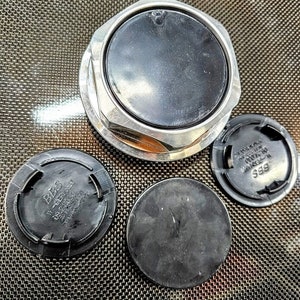 May include: Four black plastic wheel center caps with the word "BBS" printed on them. One center cap has a silver metal rim around the outside edge.