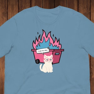 Op de afbeelding: Lichtblauw T-shirt met een cartoonkat met een bril, voor een roze vuilnisbak in brand. De vuilnisbak heeft zwarte wielen en de tekst "I can fix this" op een tekstballon.