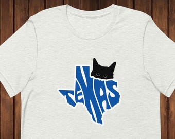 Op de afbeelding: Een lichtgrijs T-shirt met een blauwe omtrek van de staat Texas. Het woord "Texas" is binnen de omtrek gespeld. Een zwarte kat kijkt vanuit de bovenkant van de omtrek.