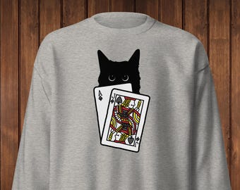 May include: A light gray sweatshirt featuring a black cat peeking over two playing cards. The cards are the Ace of Spades and the Jack of Spades. The sweatshirt is made of a soft material.