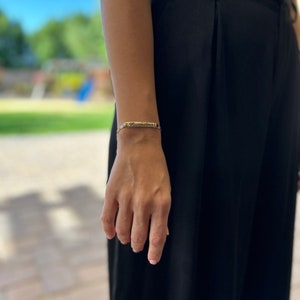 May include: A gold bracelet with a thin, rectangular charm. The charm has a textured surface and is engraved with a design.