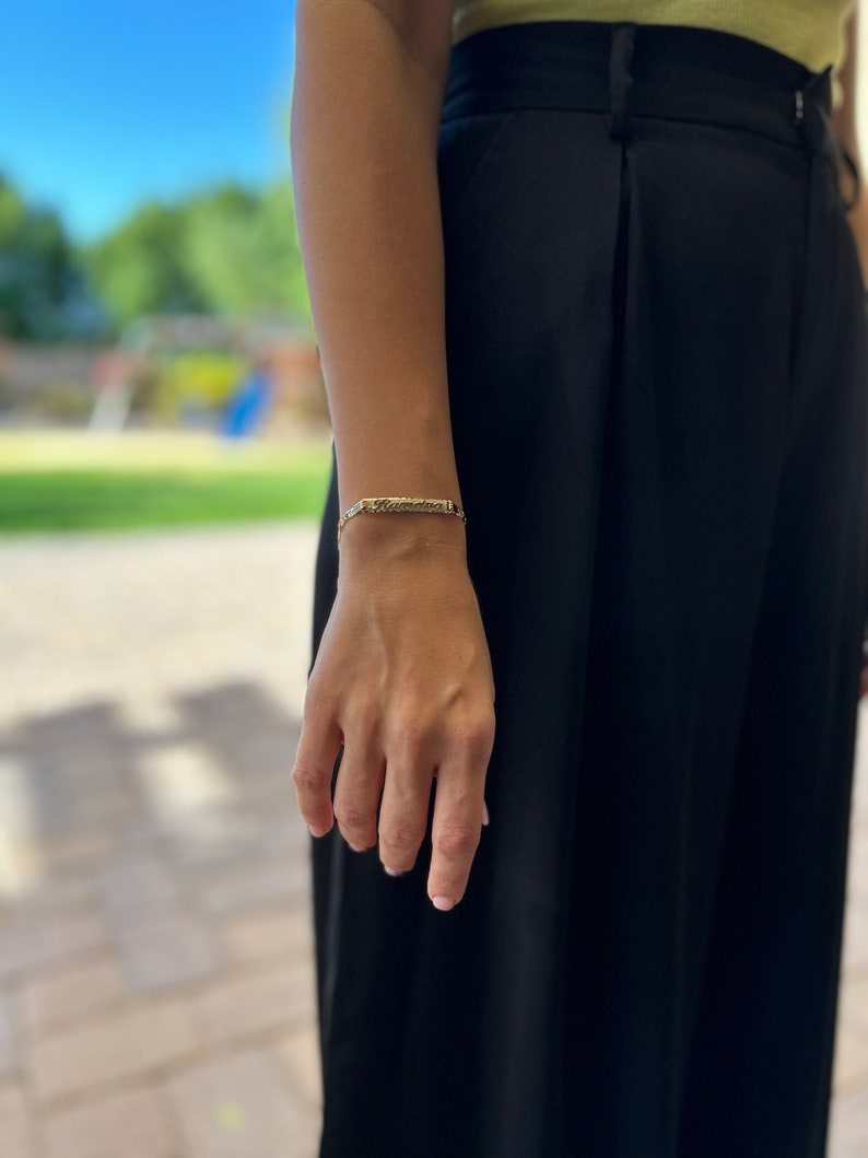 May include: A gold bracelet with a thin, rectangular charm. The charm has a textured surface and is engraved with a design.