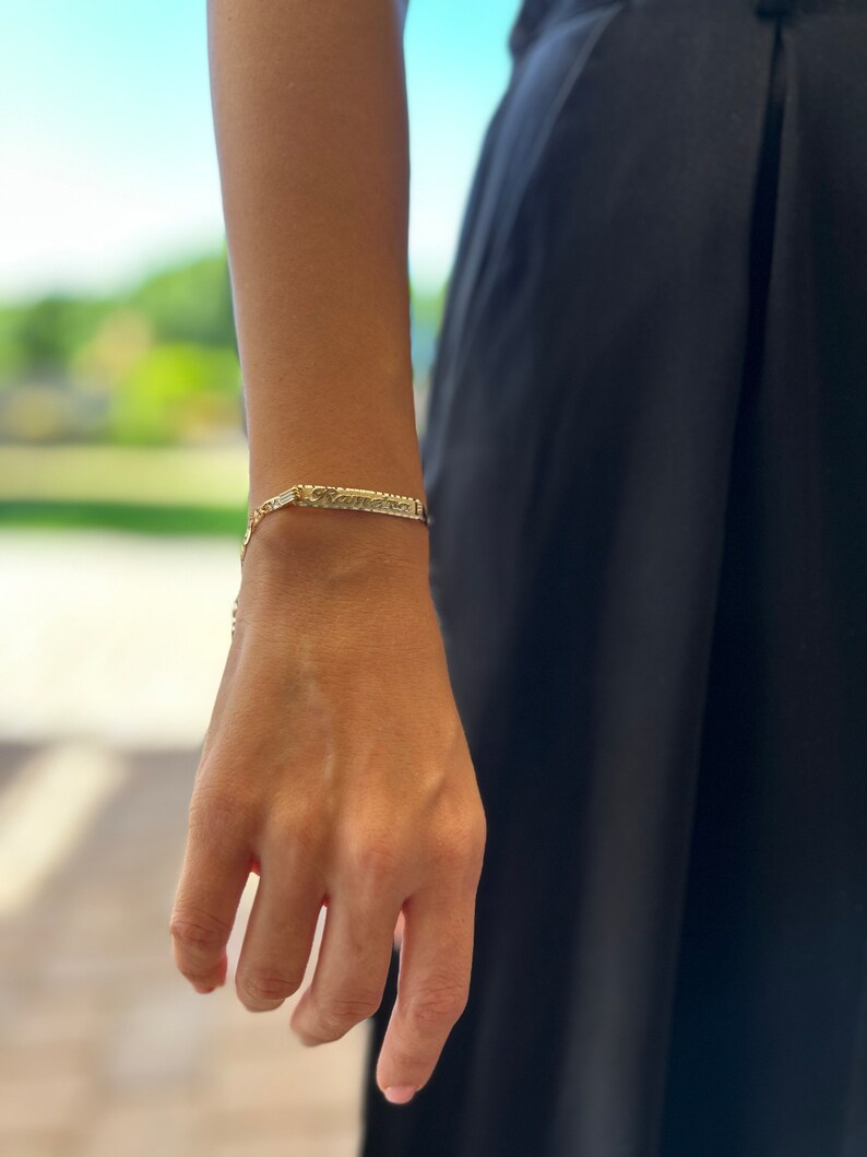 May include: A gold chain bracelet with a nameplate charm. The nameplate is engraved with the word "Kamara".