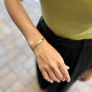 May include: A gold chain bracelet with a rectangular nameplate charm. The bracelet is on a person's wrist.
