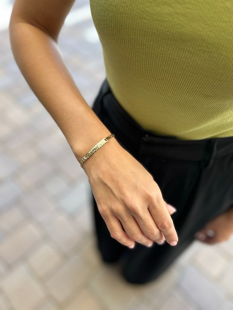 May include: A gold chain bracelet with a rectangular nameplate charm. The bracelet is on a person's wrist.
