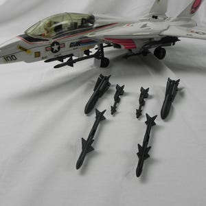 May include: A white and red toy fighter jet with black missiles. The jet has a clear cockpit and the number 100 on the side. The jet is labeled "GI Joe".