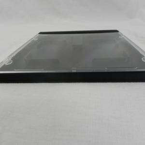 May include: A clear, rectangular CD jewel case with a black base and a transparent lid. The case is designed to hold a standard-sized compact disc. The image shows the case from a side angle, highlighting its sleek, minimalist design.