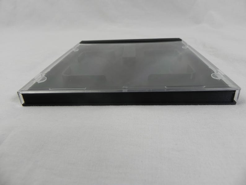 May include: A clear, rectangular CD jewel case with a black base and a transparent lid. The case is designed to hold a standard-sized compact disc. The image shows the case from a side angle, highlighting its sleek, minimalist design.
