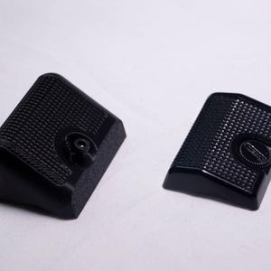 May include: Two black, textured camera grips. One grip has a rounded edge and a screw, while the other has a flat edge and a circular adjustment dial. The grips are designed for a camera and are made of a durable material.