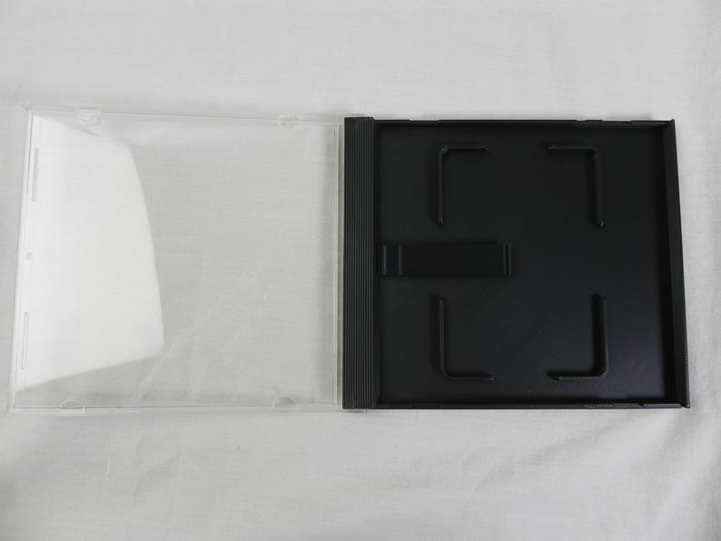 May include: An open, clear plastic CD case with a black tray. The case is rectangular with a hinged design. The black tray has a central hub and four corner clips to hold a CD in place. The case is empty.