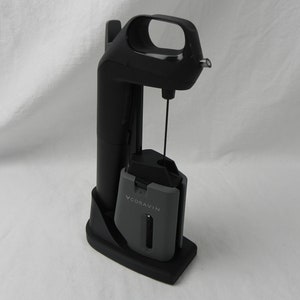 May include: A black Coravin wine system with a gray cartridge holder. The system has a tall, vertical design with a handle and a needle for inserting into a wine bottle. The gray cartridge holder has the word "CORAVIN" printed on it.