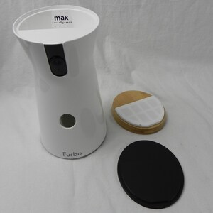 May include: A white pet camera with a black camera lens and the word "Furbo" printed on the front. The top of the device has the word "max" printed on it. Also included are a wooden lid and a black base.