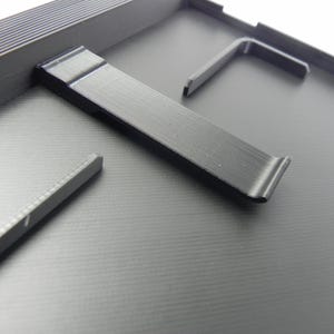 May include: Close-up of a black, textured surface with several black metal components. The components include a long, curved piece and several L-shaped pieces, possibly for assembly or support. The overall aesthetic is industrial and functional.