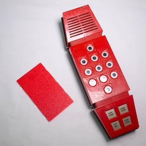 May include: A red electronic handheld game with the name "MERLIN" on the top. The game has multiple buttons and a speaker. A red rectangular piece is next to the game.