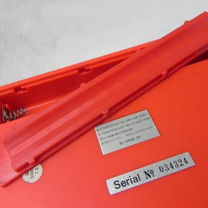 May include: A red plastic toy with a battery compartment and a silver label that reads "Serial No 034324". The label is attached to the toy with a silver screw. The battery compartment is open and the battery cover is lying next to it. The battery cover is also red plastic.