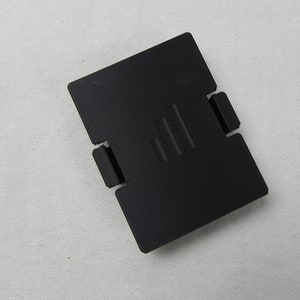 May include: Black plastic battery compartment cover with a rectangular shape and a raised design of three parallel lines.