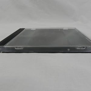 May include: A clear, rectangular CD jewel case with a black spine. The case is made of transparent plastic, allowing visibility of the contents. The case has a hinged design and is designed to hold a CD. The case is empty.