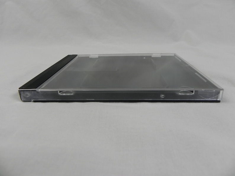 May include: A clear, rectangular CD jewel case with a black spine. The case is made of transparent plastic, allowing visibility of the contents. The case has a hinged design and is designed to hold a CD. The case is empty.