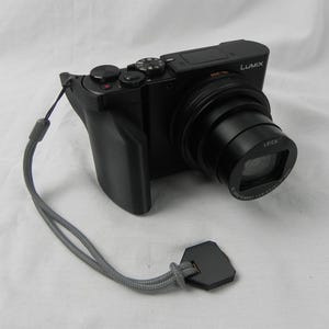 May include: A black digital camera with a lens and a strap. The camera has a silver button and the word "LUMIX" printed on the side.