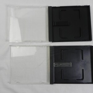 May include: Two clear plastic CD cases with black trays. The cases are open, showing the clear plastic front and the black tray with a central locking mechanism. The cases are on a white surface.