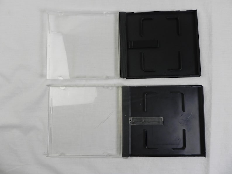 May include: Two clear plastic CD cases with black trays. The cases are open, showing the clear plastic front and the black tray with a central locking mechanism. The cases are on a white surface.
