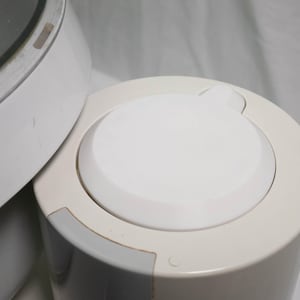 Peut inclure: Gros plan d'un appareil blanc avec un dessus arrondi et un couvercle circulaire. Le couvercle a une petite poignée incurvée. L'appareil est cylindrique et a une surface lisse et propre. Le fond est blanc uni.