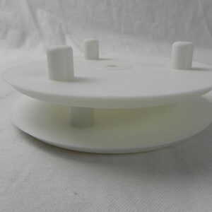 May include: White plastic spool with four raised knobs on the top and bottom. The spool is designed to hold thread or yarn.