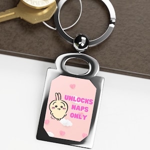 May include: A silver keychain with a rectangular charm featuring a cartoon bunny and the text "UNLOCKS NAPS ONLY" on a pink background. The keychain is attached to a keyring with two gold keys.