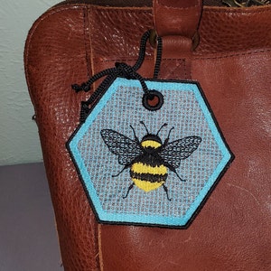 May include: A blue and white hexagonal tag with a black and yellow embroidered bee design. The tag is attached to a brown leather bag.