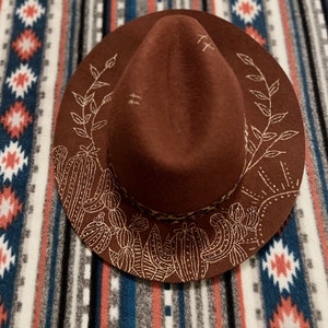 May include: A brown felt fedora hat with a wide brim, featuring a gold embroidered design of cacti and leaves. The hat has a brown leather band with a leopard print pattern.