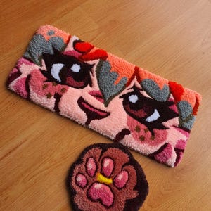 Könnte beinhalten: Ein rechteckiger getufteter Teppich mit einem stilisierten Anime-Gesicht mit großen dunklen Augen, rosafarbenen Hauttönen und orangefarbenem Haar, begleitet von einem kleineren runden getufteten Teppich in Form einer dunkelbraunen Tierpfote mit rosa und gelben Ballen.