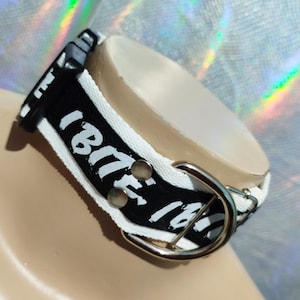Könnte beinhalten: Ein schwarz-weißes Choker-Halsband mit dem Text "I BITE 16" in Weiß. Das Halsband hat eine silberfarbene Schnalle und zwei silberfarbene Nieten. Das Halsband befindet sich auf einer beigen Oberfläche.