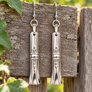 May include: A pair of silver-toned drop earrings featuring a long, rectangular metal design with embossed floral and scrollwork details. Each earring hangs from a simple silver-toned hook wire, showcasing a polished metallic finish and a vertical, tapered shape with intricate decorative patterns along the center.