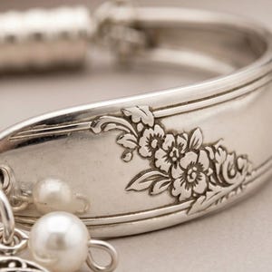 May include: A silver-toned metal cuff bracelet crafted from a repurposed spoon handle, featuring an embossed floral pattern along the outer surface. The bracelet is adorned with two small, round, white faux pearl beads hanging from a metal jump ring.