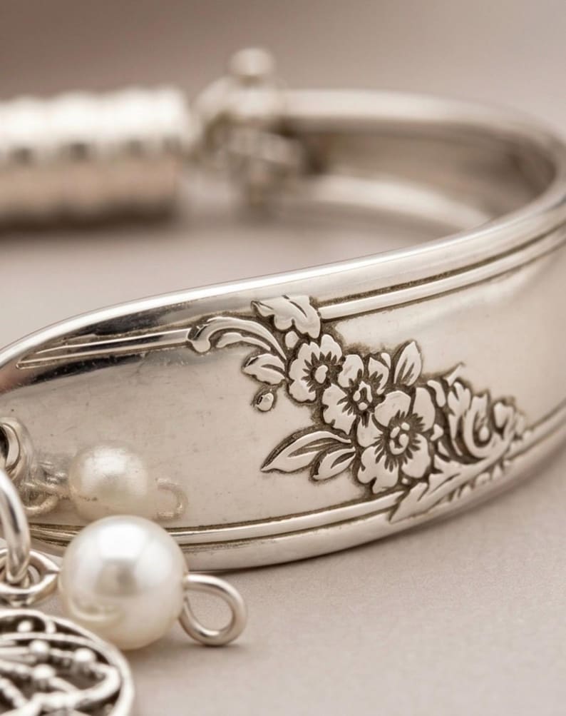 May include: A silver-toned metal cuff bracelet crafted from a repurposed spoon handle, featuring an embossed floral pattern along the outer surface. The bracelet is adorned with two small, round, white faux pearl beads hanging from a metal jump ring.