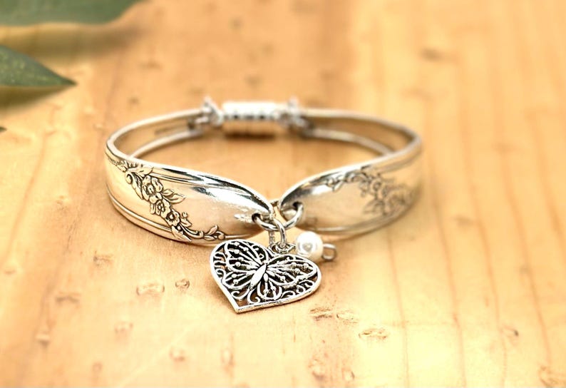 May include: A silver-toned spoon bracelet featuring an embossed floral pattern along the band. The bracelet includes a dangling heart-shaped charm with an intricate butterfly cutout design and a small white bead accent, secured with a magnetic clasp.