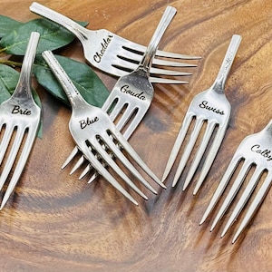May include: Six silver cheese forks with engraved cheese names: Brie, Blue, Cheddar, Gouda, Swiss, and Colby. The forks are arranged on a wooden surface with green leaves. A unique and elegant addition to any cheese board.