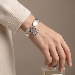May include: A silver-toned bracelet crafted from repurposed spoon handles featuring an intricate floral pattern. The bracelet includes a dangling heart-shaped charm with a detailed butterfly cutout design and a small, round white pearl accent.