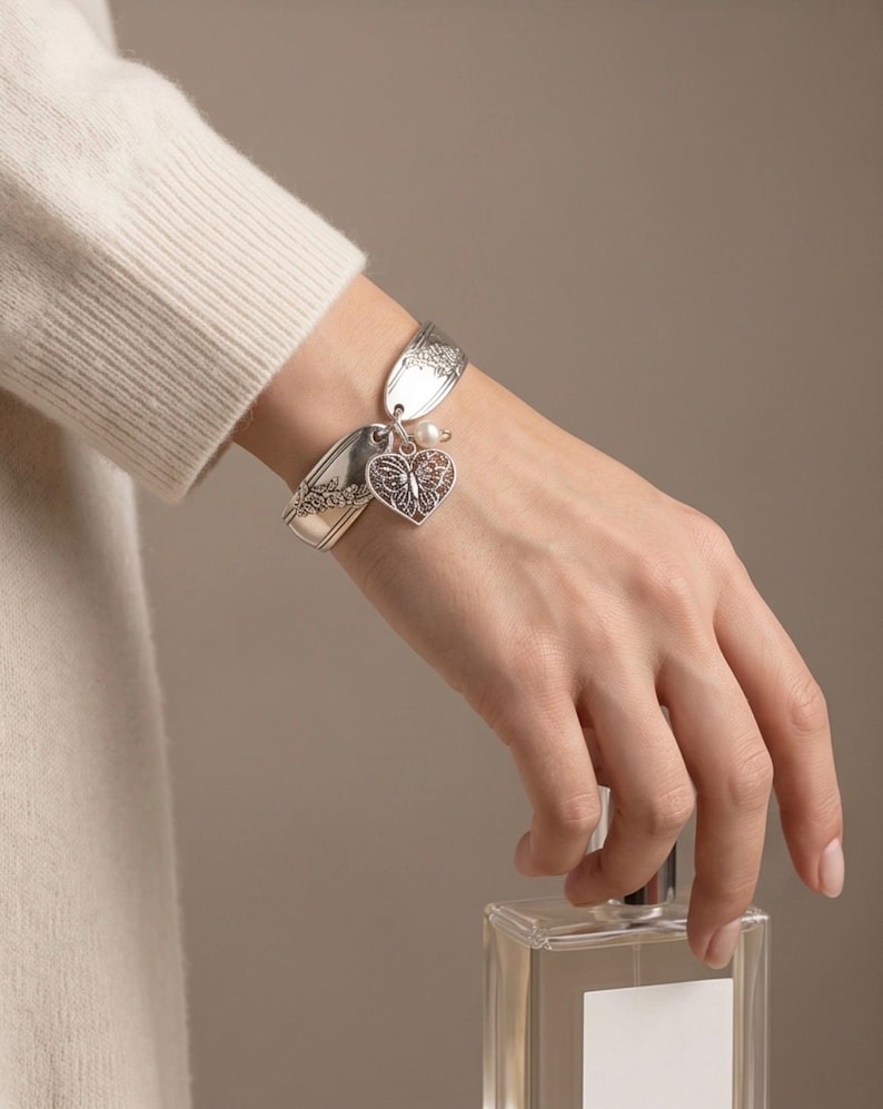 May include: A silver-toned bracelet crafted from repurposed spoon handles featuring an intricate floral pattern. The bracelet includes a dangling heart-shaped charm with a detailed butterfly cutout design and a small, round white pearl accent.