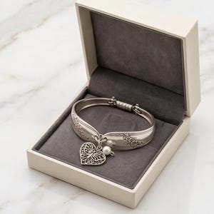 May include: A silver-toned hinged bracelet crafted from a vintage spoon handle, featuring an embossed floral pattern. A heart-shaped charm with an intricate filigree butterfly design and a small white pearl bead hang from the center of the bracelet.