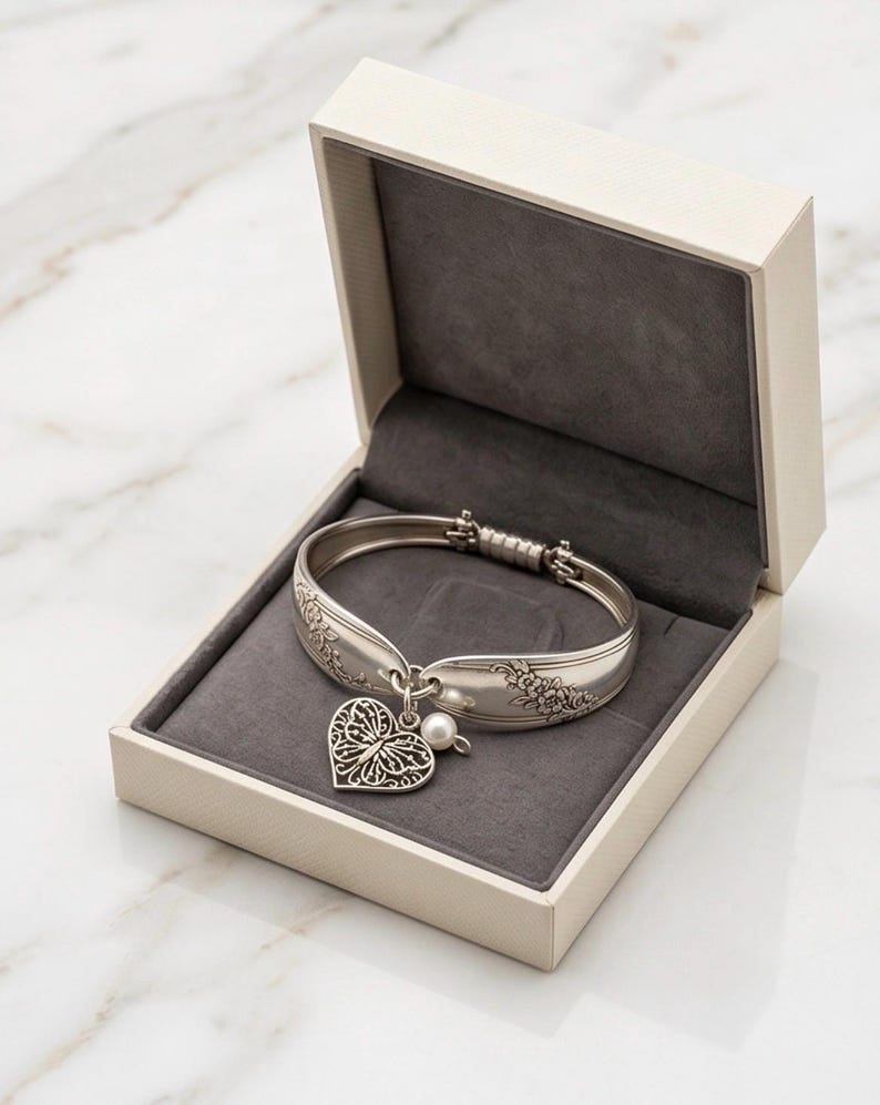 May include: A silver-toned hinged bracelet crafted from a vintage spoon handle, featuring an embossed floral pattern. A heart-shaped charm with an intricate filigree butterfly design and a small white pearl bead hang from the center of the bracelet.