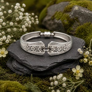 May include: A silver-toned metal cuff bracelet crafted from two spoon handles joined by a central ring. The bracelet features an embossed floral pattern along the outer surface and a cylindrical magnetic clasp at the opening for closure.