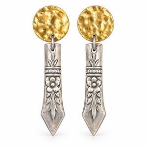May include: A pair of drop earrings featuring a circular, hammered gold-tone top piece connected to a long, silver-tone rectangular pendant. The silver pendant displays an embossed floral motif with leaves and a central flower design.