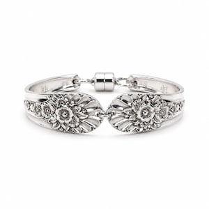 May include: A silver-toned bracelet crafted from two curved spoon handles featuring an intricate embossed floral pattern. The two ends are connected by a cylindrical magnetic clasp, creating a continuous loop with a polished metallic finish.